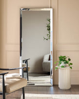 Large rectangular leaner mirror with silver frame, displayed against beige wall next to potted plant and chair.