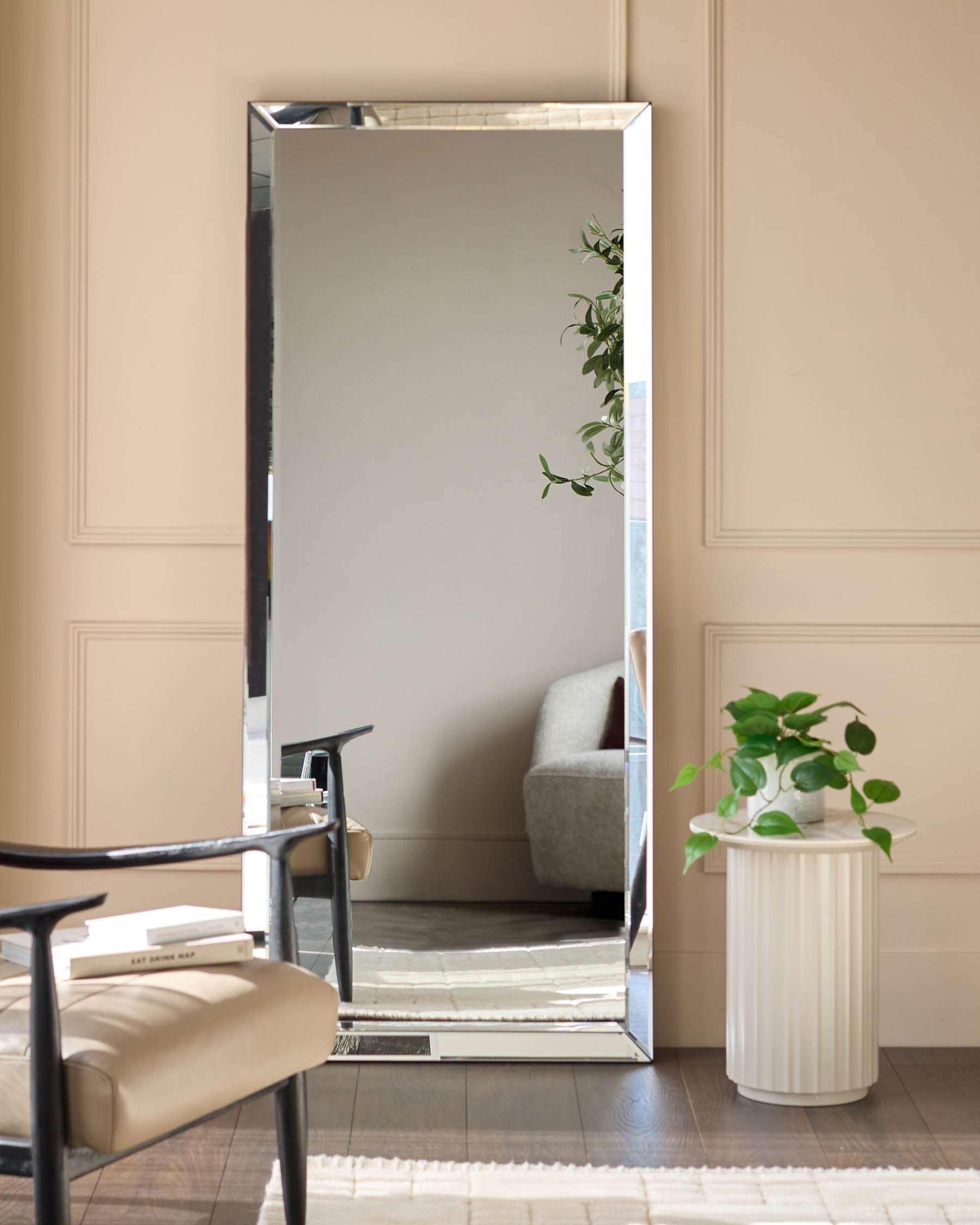 Large rectangular leaner mirror with silver frame, displayed against beige wall next to potted plant and chair.