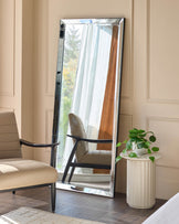 Large rectangular leaner mirror with polished silver frame, displayed against beige wall next to black chair and potted plant.