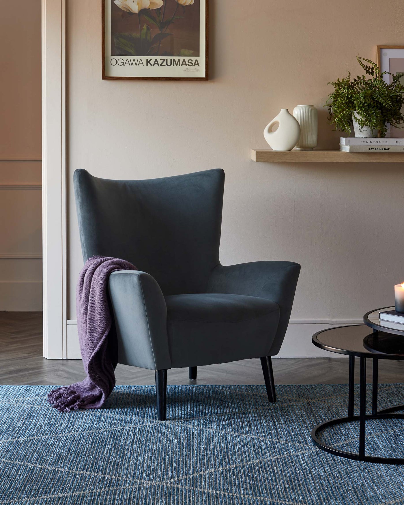Elegant modern armchair with a high, curvilinear backrest and plush seating, upholstered in a smooth, dark grey fabric. It features sleek, tapered black legs. Next to it is a small, round, black metal side table with a glass top. A pale purple throw blanket is casually draped over one arm of the chair. The furniture sits on a textured blue area rug, adding a pop of colour to the neutral-toned room.