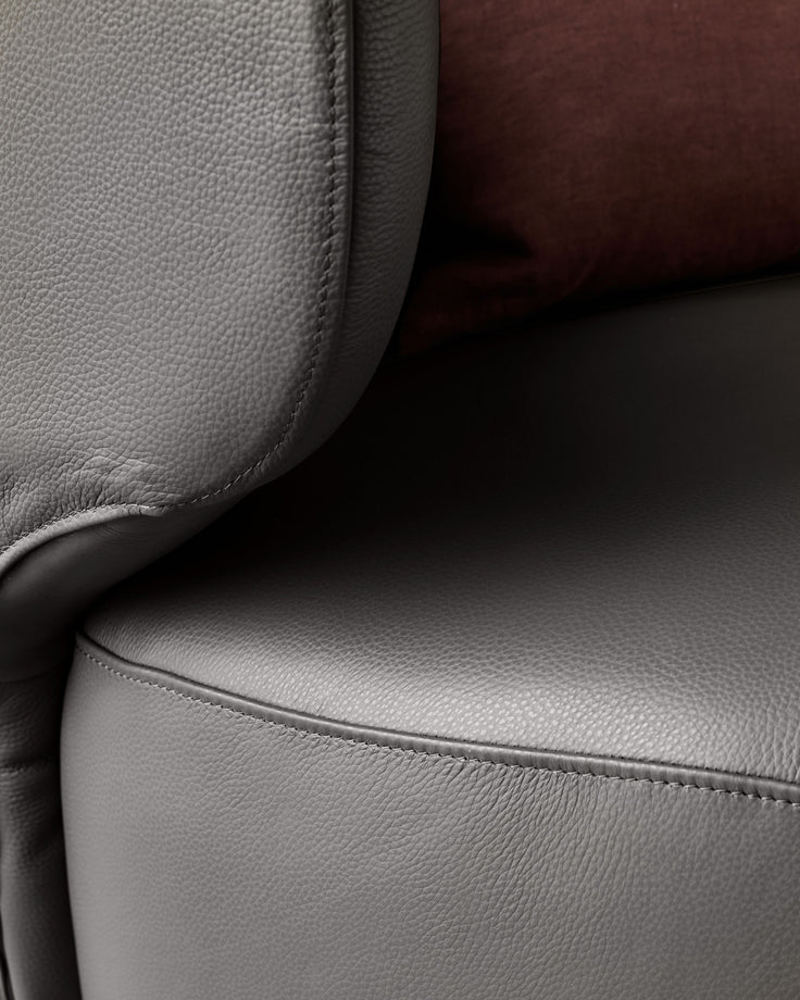 Close-up of a grey leather sofa, showing texture and stitching. A brown cushion rests in the background.
