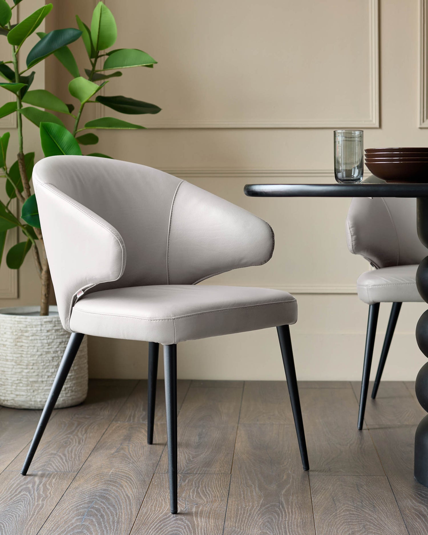 Light grey leather dining chair with curved backrest and black tapered legs, shown in styled room setting.