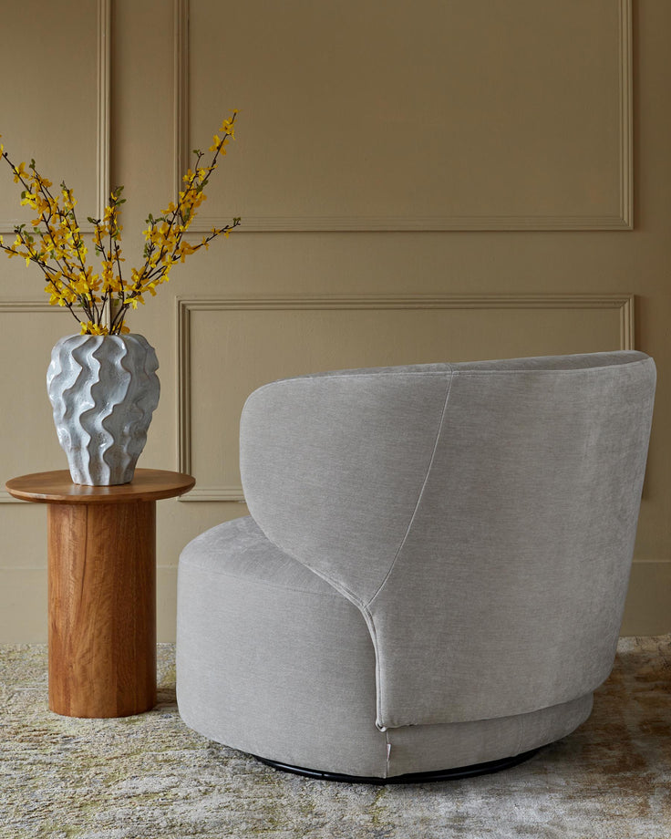 A modern grey swivel chair with a wooden side table and flower arrangement against a neutral wall.