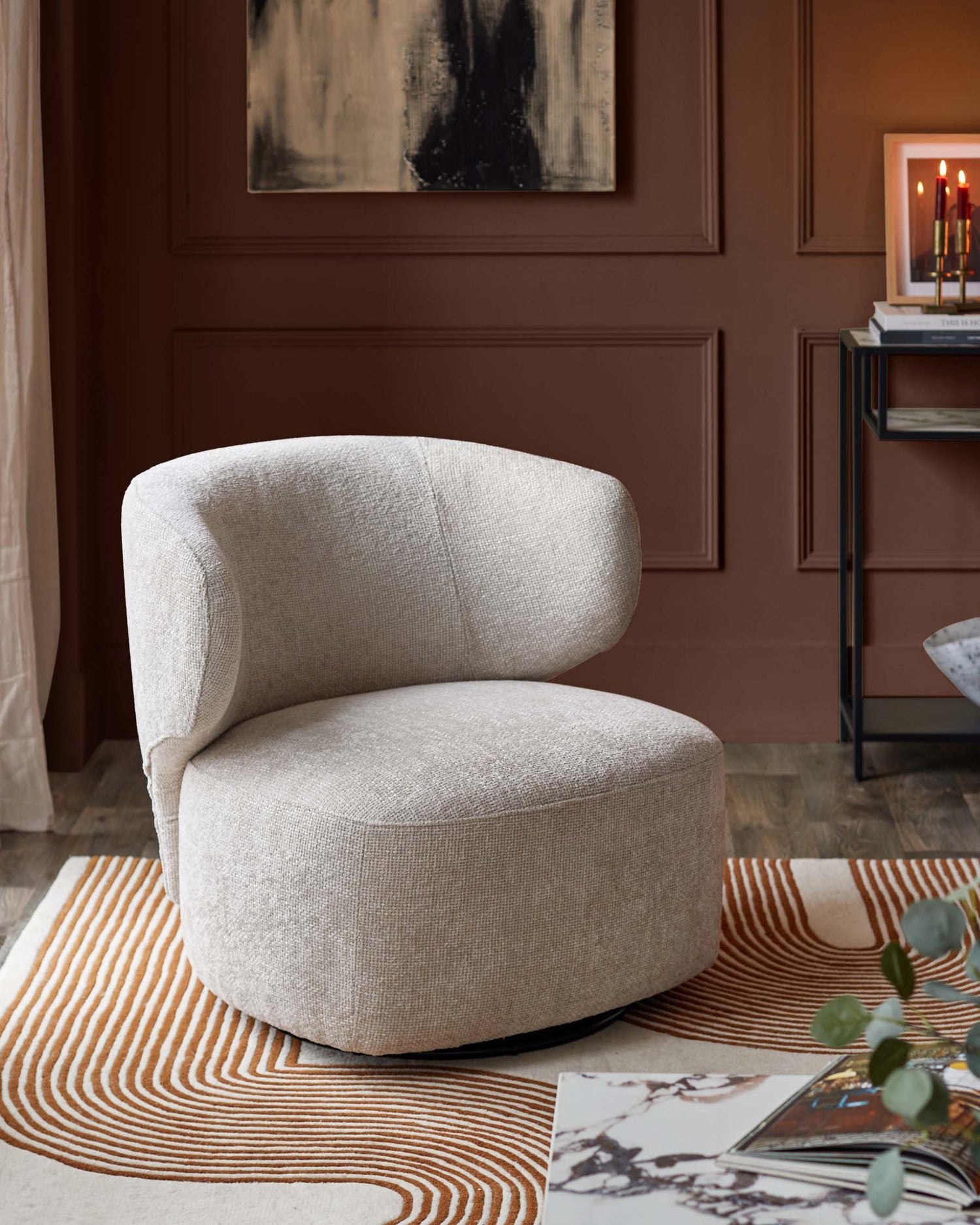 Stylish cream-coloured swivel chair sits on a patterned rug, beside a dark brown wall with art and a side table. Modern interior design.