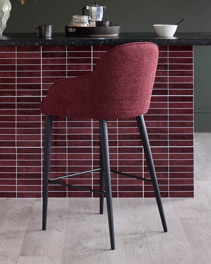 A modern bar stool with a plush, red fabric seat and sleek black legs, positioned in front of a tiled bar area.