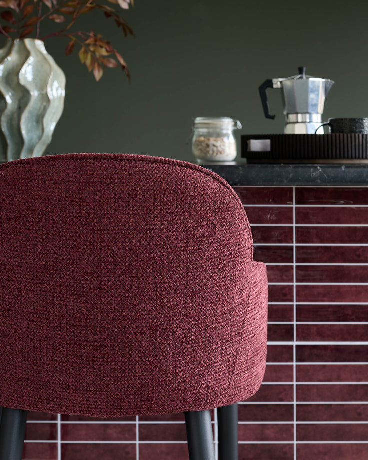 Burgundy chair in front of a kitchen island with accessories such as coffee maker and a jar of oats.