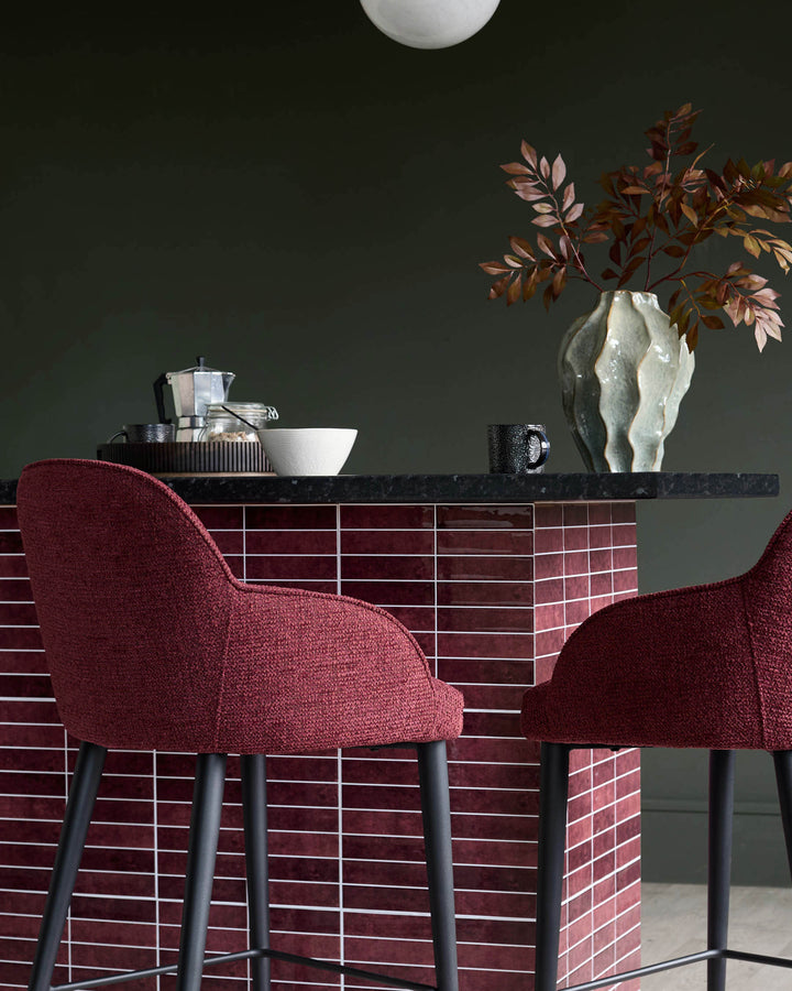 Two red bar stools sit at a tiled counter with a black marble top, displaying coffee & ornaments.
