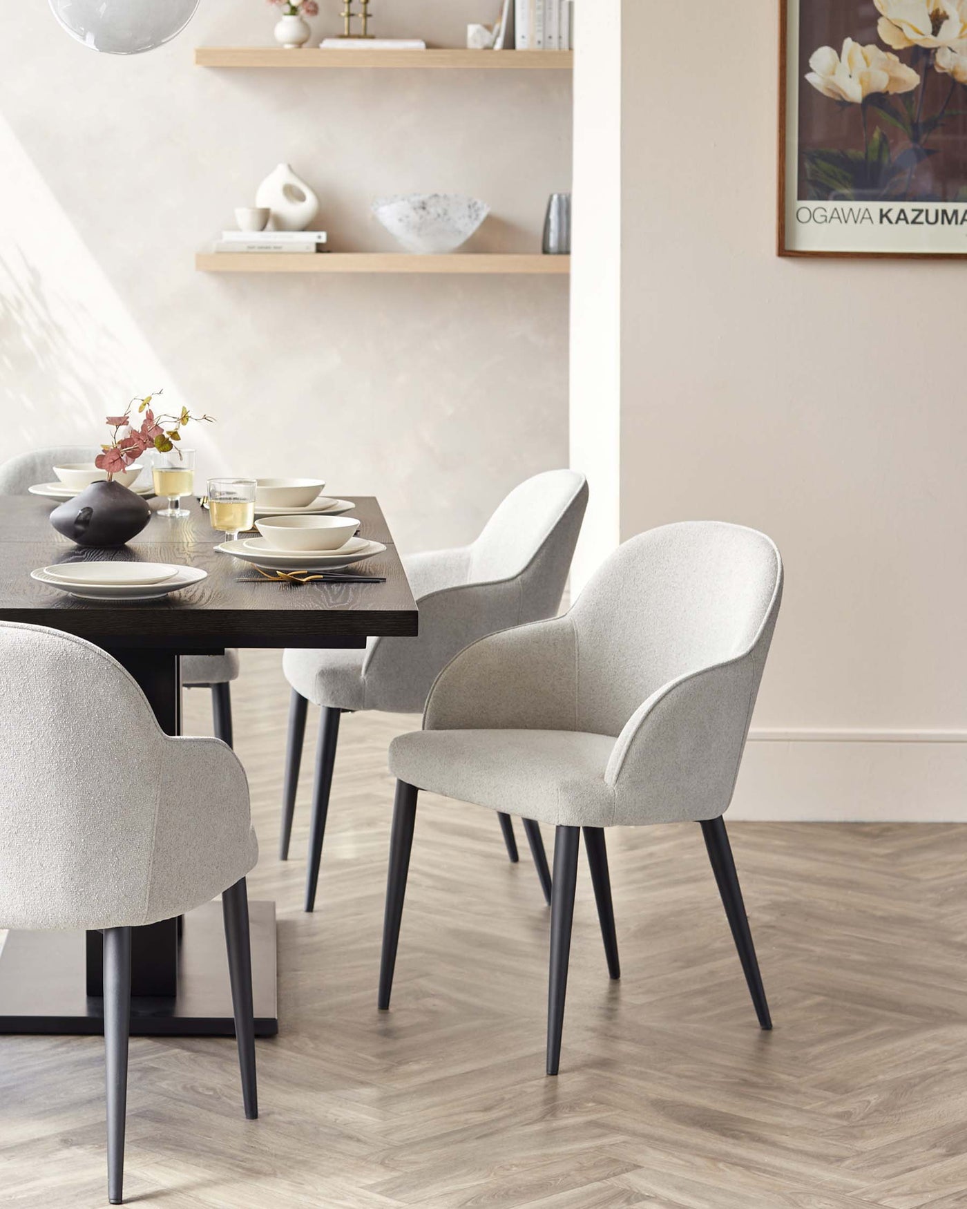 A contemporary dining set featuring a dark wooden table with a textured finish and angled, black legs. Accompanied by three light grey upholstered chairs with a smooth, curved design and slender black legs that coordinate with the table. The chairs have a slightly winged backrest and armless construction, providing a modern yet comfortable seating experience. The table is set with simple white and dark plates, bowls, and glassware.