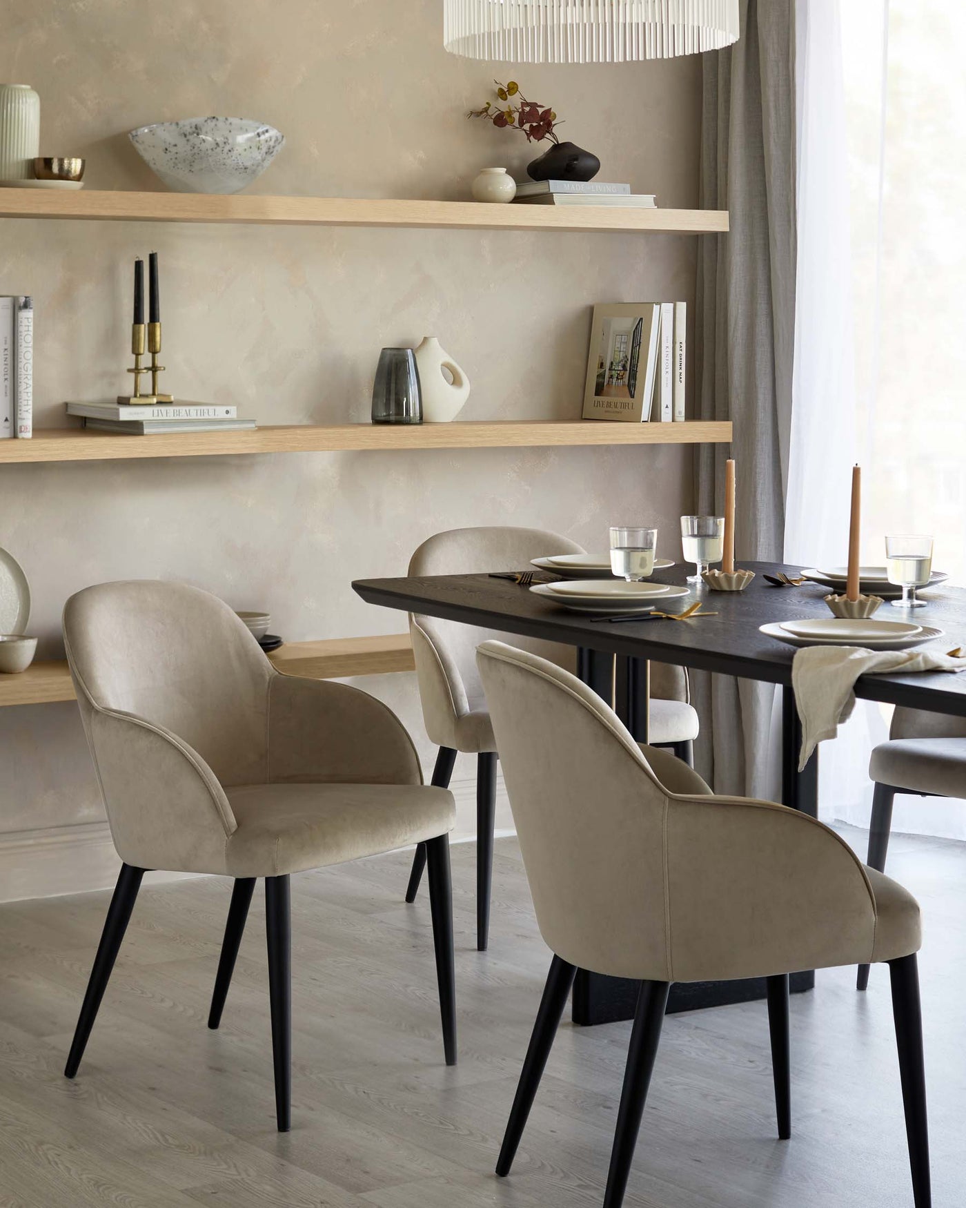 A modern dining area featuring an oval-shaped dark wood table with a matte finish and black tapered legs, surrounded by four elegant upholstered chairs with a plush beige fabric. The chairs showcase a curvaceous silhouette with soft lines and black tapered legs to match the table, offering a sophisticated and cohesive design. On the wall, two natural wood floating shelves are placed, storing decorative items and books, contributing to the minimalistic yet warm ambiance of the space.
