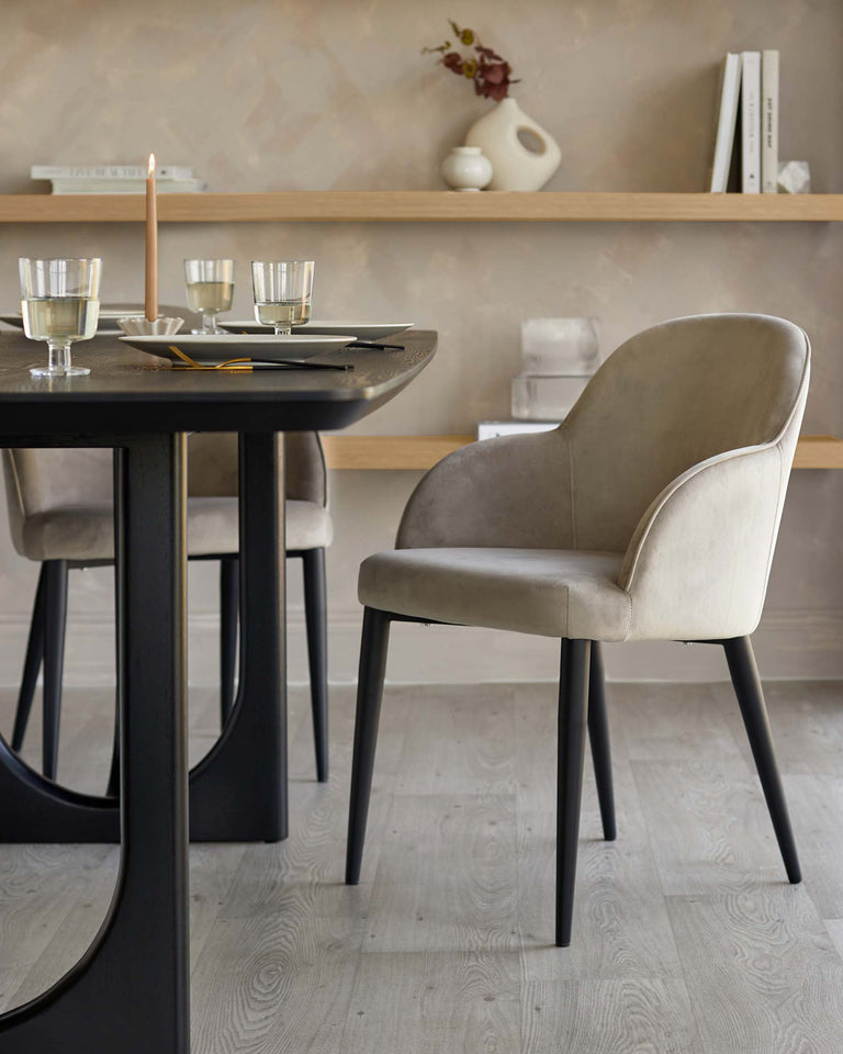 A modern round dining table with a black finish and a set of elegant taupe upholstered dining chairs with black legs. The table is set with plates, glasses, and candles, suggesting an intimate dining setting. A floating shelf with decor items is visible in the background, complementing the minimalist and chic arrangement.