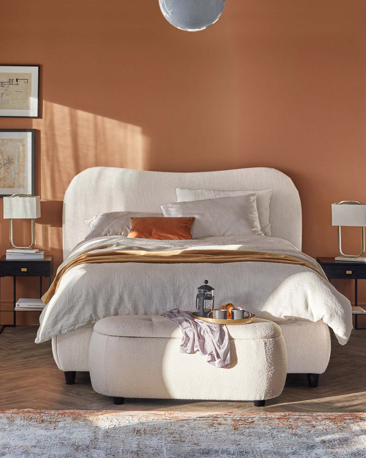 Cozy bed with a soft, curved headboard, plush ottoman, modern bedside lamps, and warm, textured bedding against an earthy backdrop.
