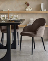 Mocha velvet dining chair with curved backrest and black legs, shown at round black table in dining room setting.