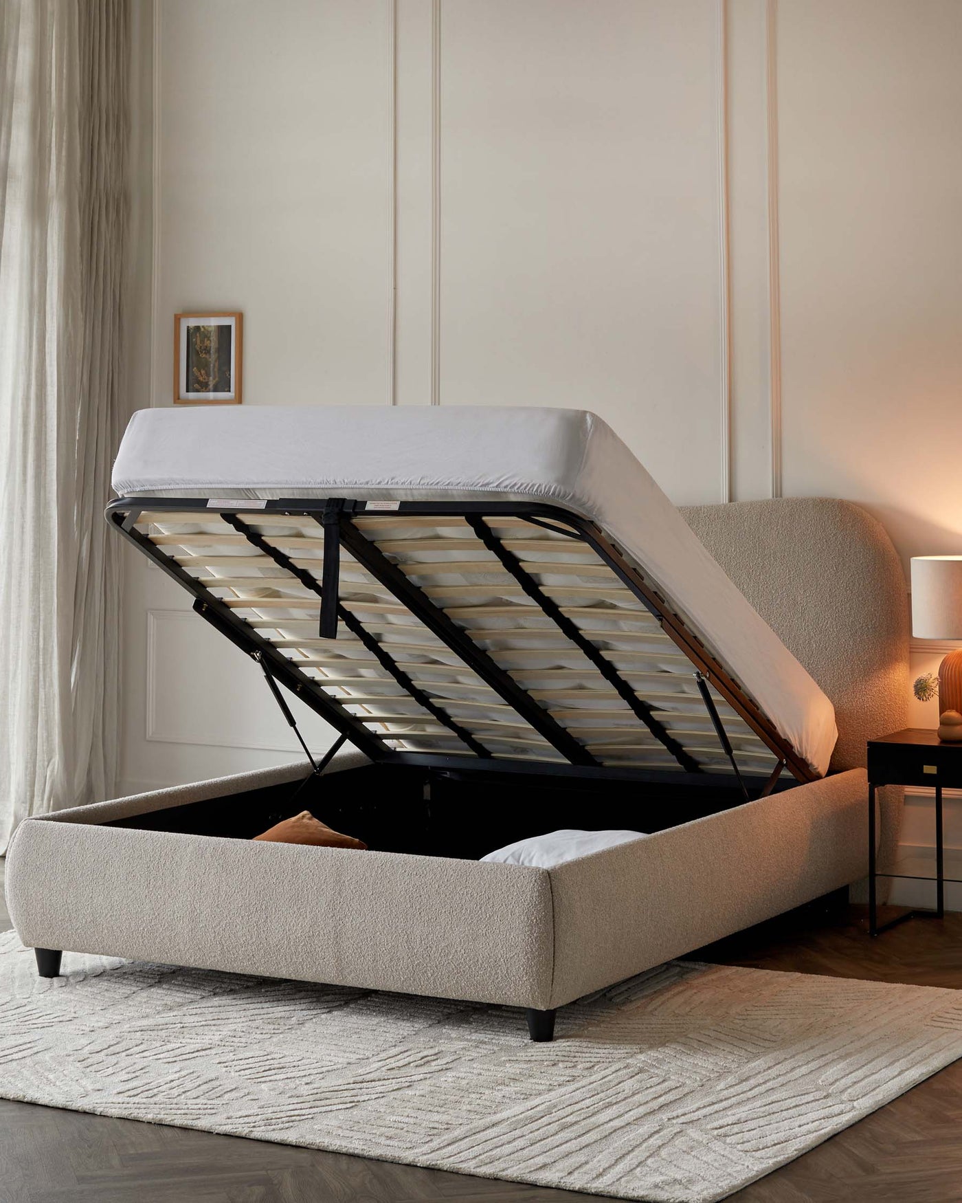 Upholstered storage bed with hydraulic lift system, featuring a beige fabric frame on black legs, a lifted mattress revealing an open storage space, set on a textured area rug, accented by a warm-glow table lamp and beside table.