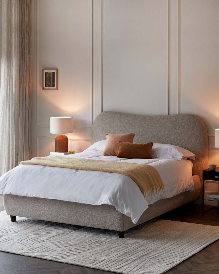 A modern upholstered queen-sized bed in a neutral colour with a gently curved headboard and tapered black legs. On either side of the bed are two minimalist white bedside tables, each with a single drawer and an open shelf beneath. Atop each table is a modern lamp with a cylindrical white shade and a base of warm wood tones. The ensemble is complemented by a textured grey area rug beneath the bed.