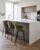 Lani bar stool in olive green velvet with black metal legs, shown at kitchen island with marble countertop.