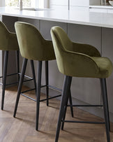 Lani bar stools in olive green velvet with black wooden legs, positioned at a white kitchen island counter.