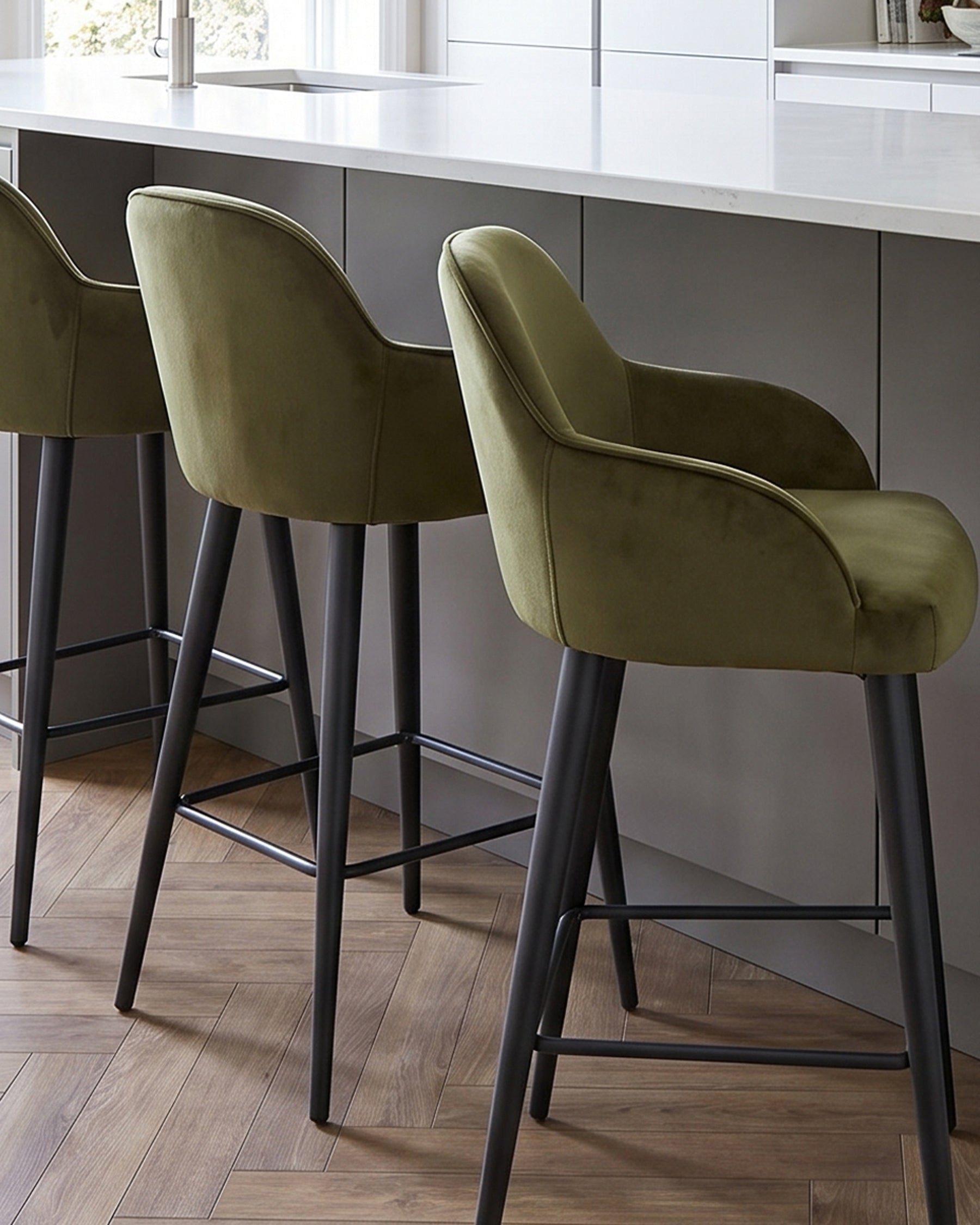 Lani bar stools in olive green velvet with black wooden legs, positioned at a white kitchen island counter.