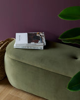 A stylish, olive-green, velvet ottoman with button detailing, topped with books, against a rich plum backdrop.