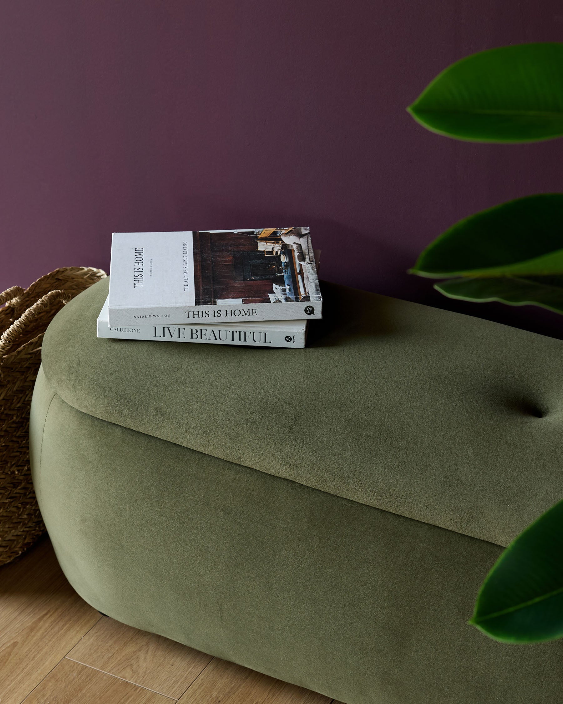 A stylish, olive-green, velvet ottoman with button detailing, topped with books, against a rich plum backdrop.