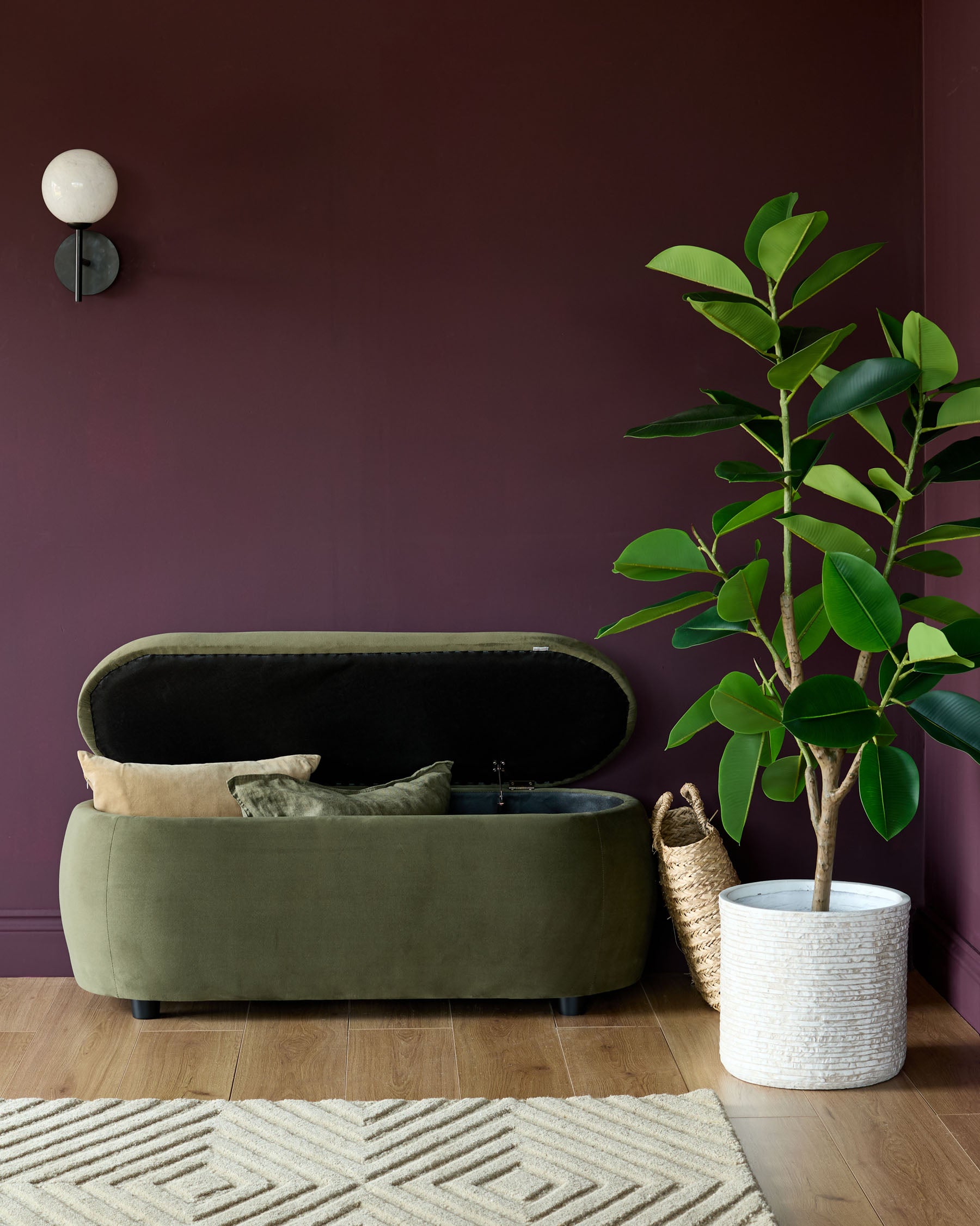 Olive ottoman storage bench with lid open next to a rubber plant in a textured white pot on a wooden floor.