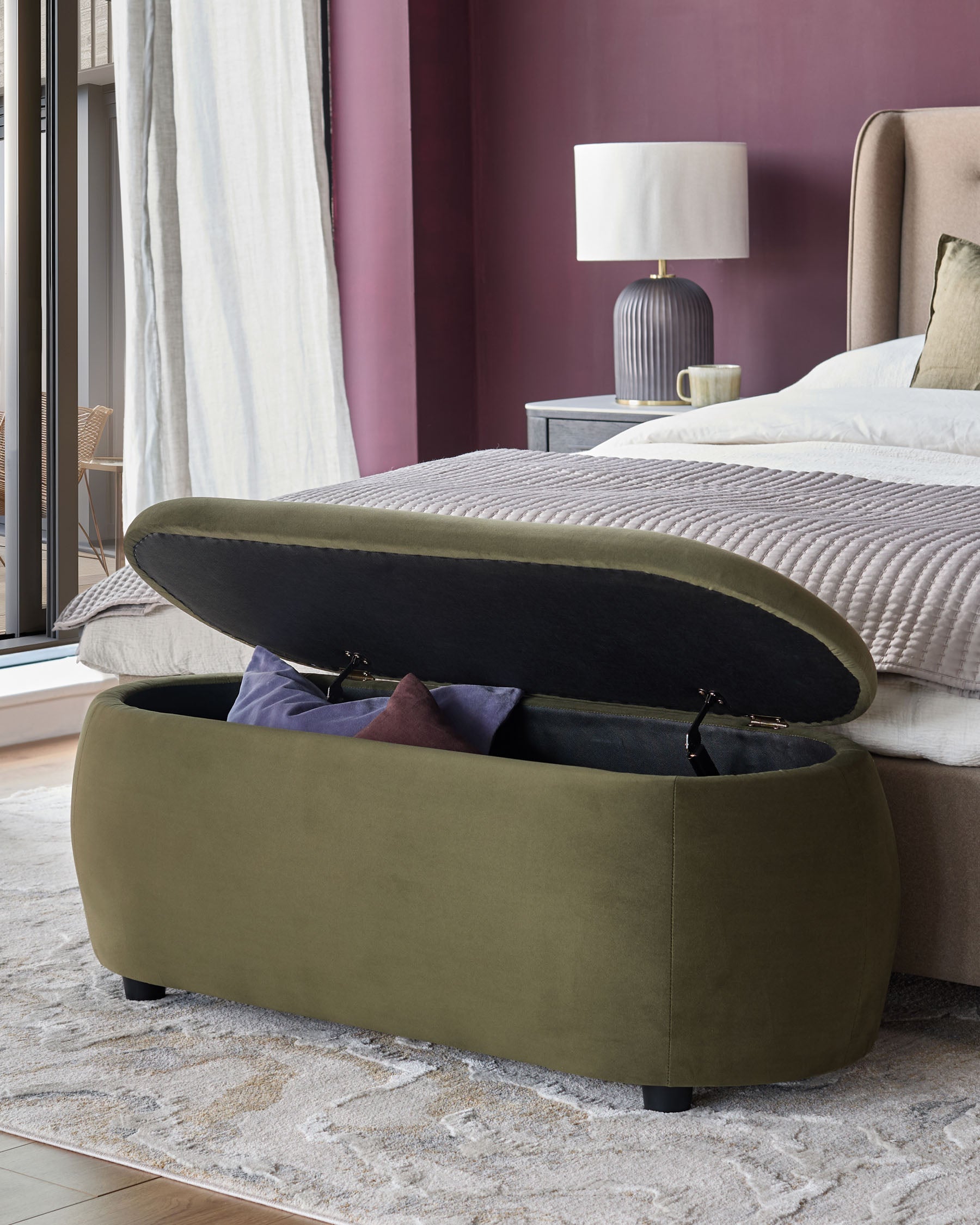 An olive green storage ottoman is open at the end of a bed, revealing pillows inside.