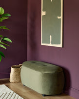 Olive green storage ottoman, a seagrass basket and modern artwork, on a wooden floor against a plum wall.