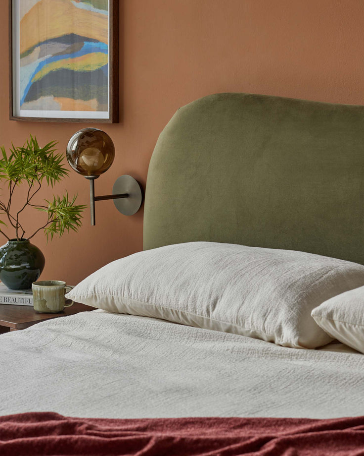 Bedroom featuring an olive headboard, a bedside table with a lamp and plant, and a bed with a cream duvet and a pink throw.