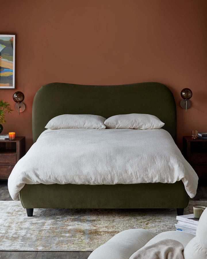 Olive green upholstered bed with a curved headboard, bedside tables with lamps, and a patterned rug on a wooden floor.