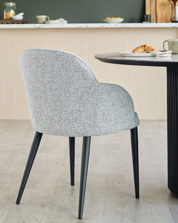 Upholstered chair with black legs next to a round table with a croissant and a mug on it.