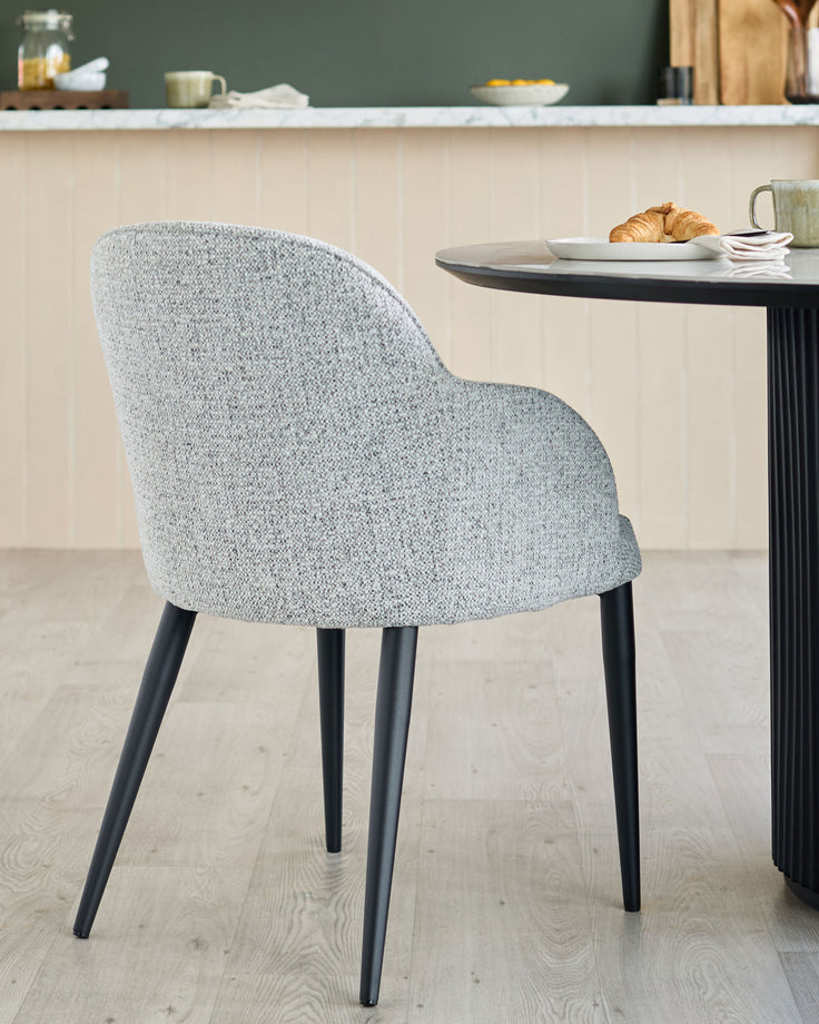 Upholstered chair with black legs next to a round table with a croissant and a mug on it.