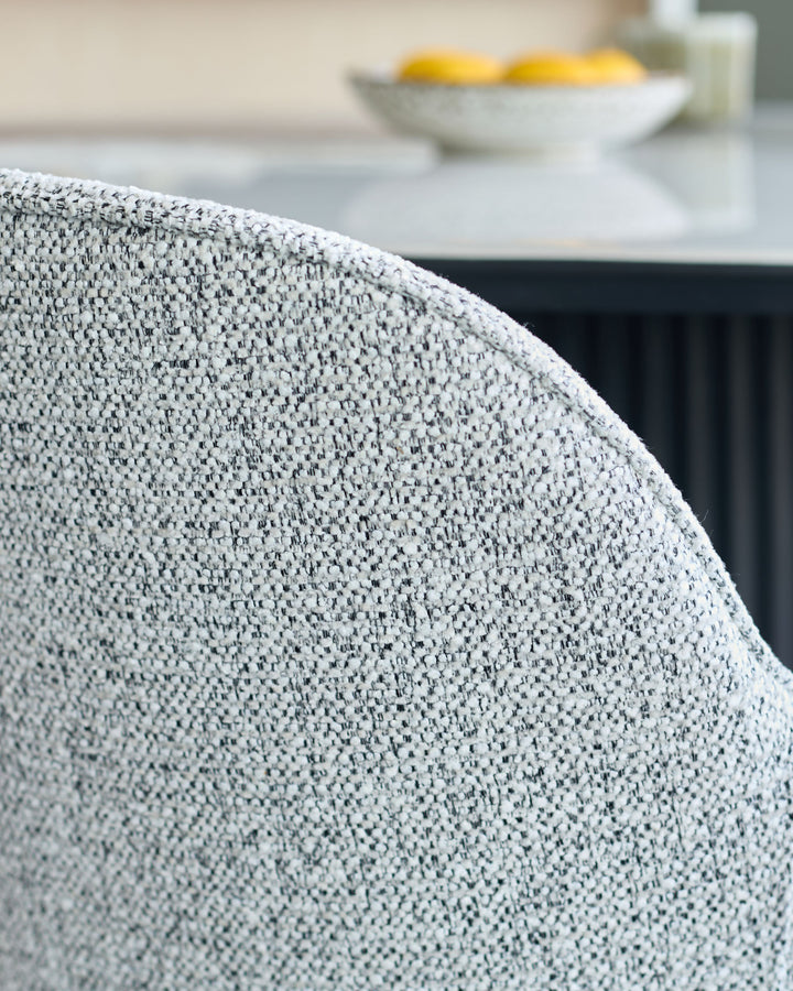 Close-up of grey textured chair back. Dining table and fruit bowl in background.