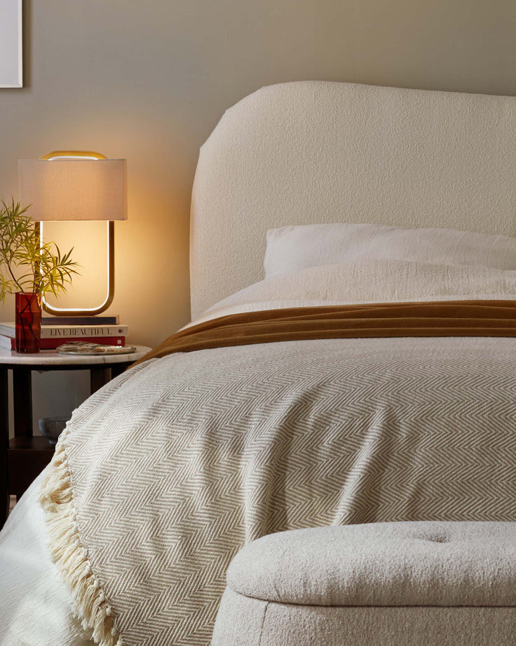 Bedroom scene with upholstered headboard, patterned blanket, and side table with lamp and decorative items.