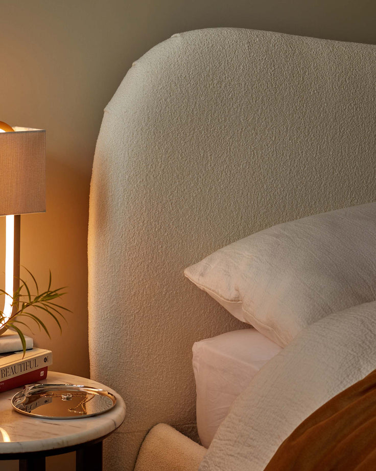 A close up of a bed with a light coloured, upholstered headboard, next to a lamp and marble side table.