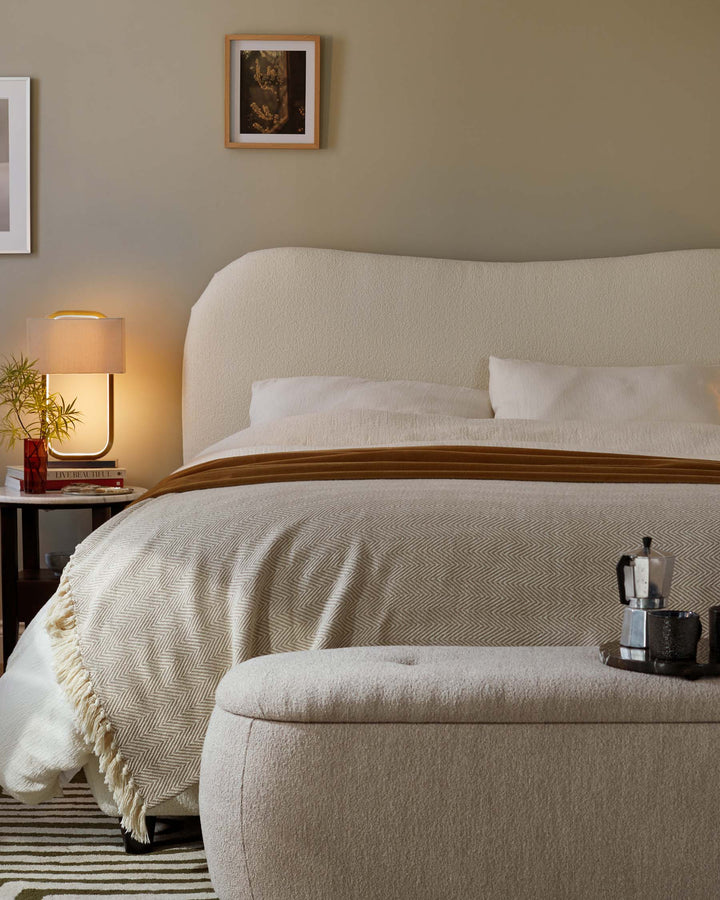 Bedroom with off-white upholstered bed, ottoman & bedside table with lamp, vase of flowers and books.