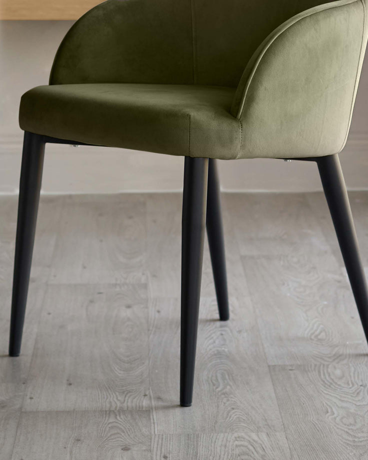 Lani dining chair in olive green velvet with curved backrest and black tapered legs, shown from side angle on light wood flooring.