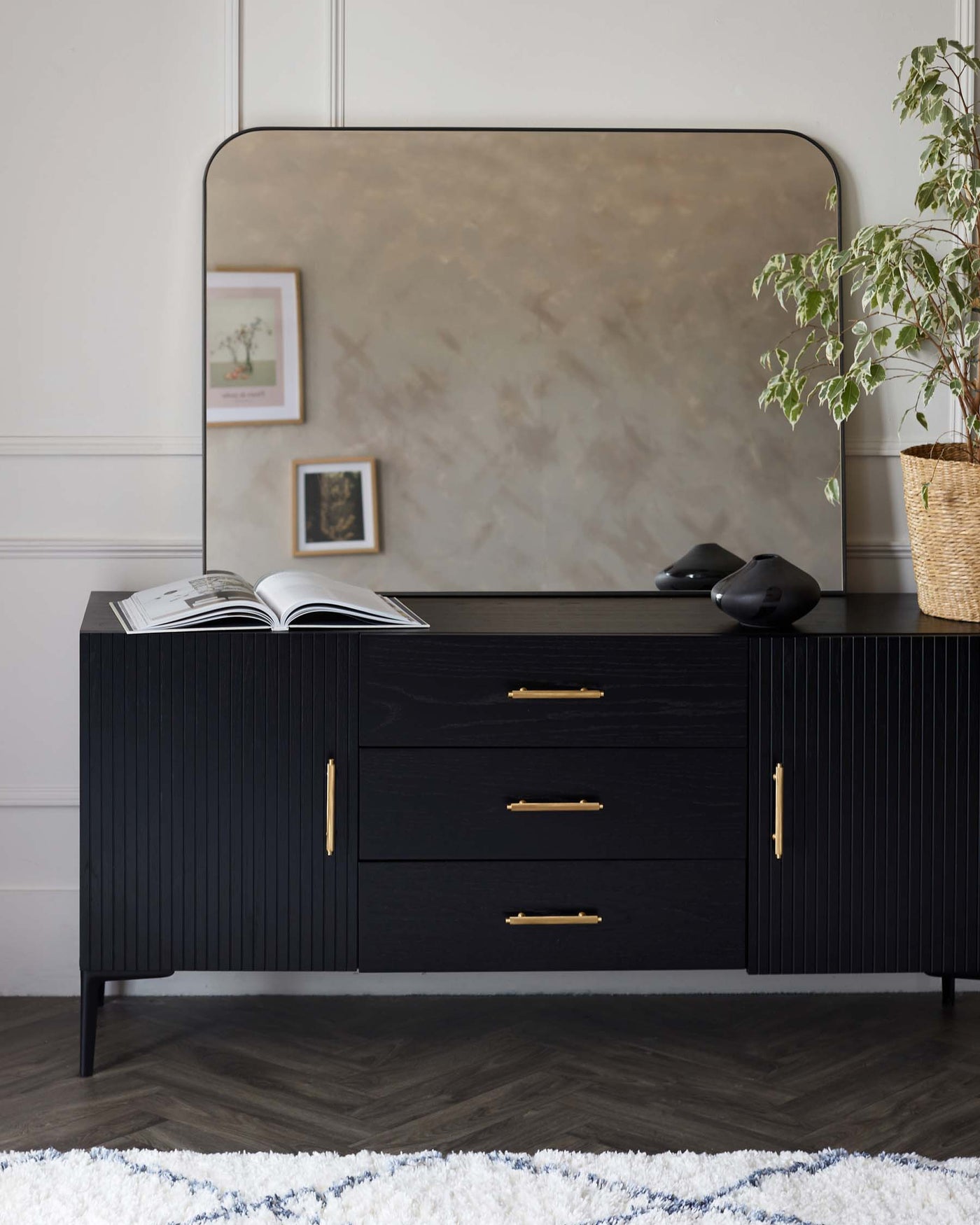 Elegant black wooden sideboard with ribbed detailing and brass hardware, accompanied by a large, rectangular, slightly curved, smoke-tinted mirror. The sideboard features three drawers and two side cabinets, set against a neutral-toned wall above a textured white and blue area rug. Decor includes a potted indoor plant, decorative black sculptures, and a laid-open book.