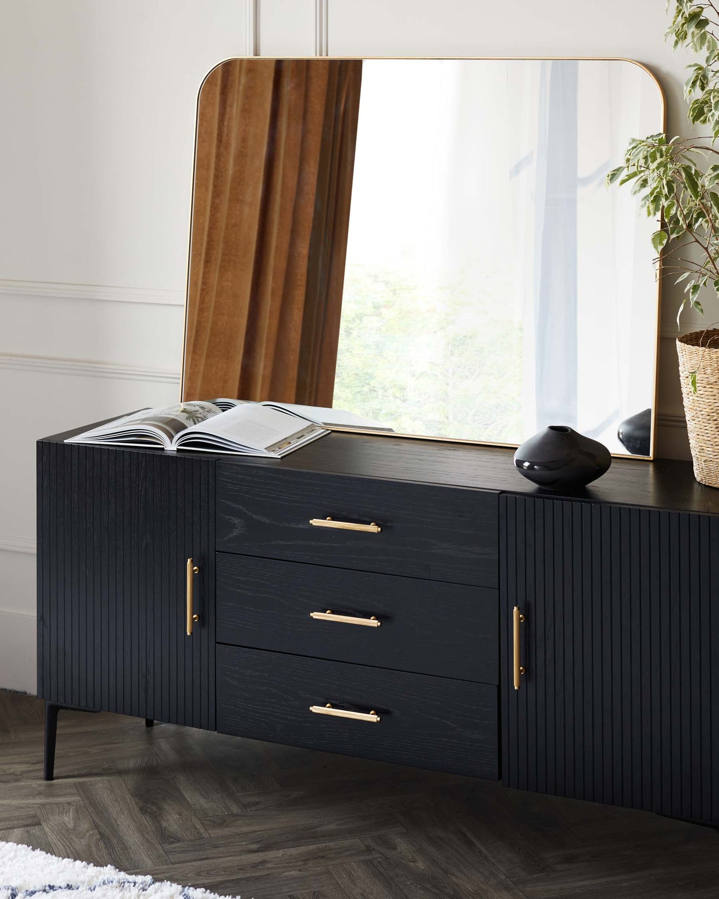 A contemporary black wooden sideboard with ribbed front panels and brass handles, paired with a large rectangular mirror featuring a rounded top and brass-coloured frame. The sideboard has three drawers with brass pull handles and stands on slender tapered legs. A decoratively placed open book, a small black vase, and a woven basket with greenery are arranged to accent the piece.