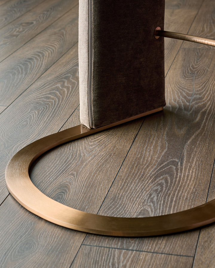Leighton brushed bronze bar stool base detail showing curved footrest and mocha fabric seat on wood flooring.