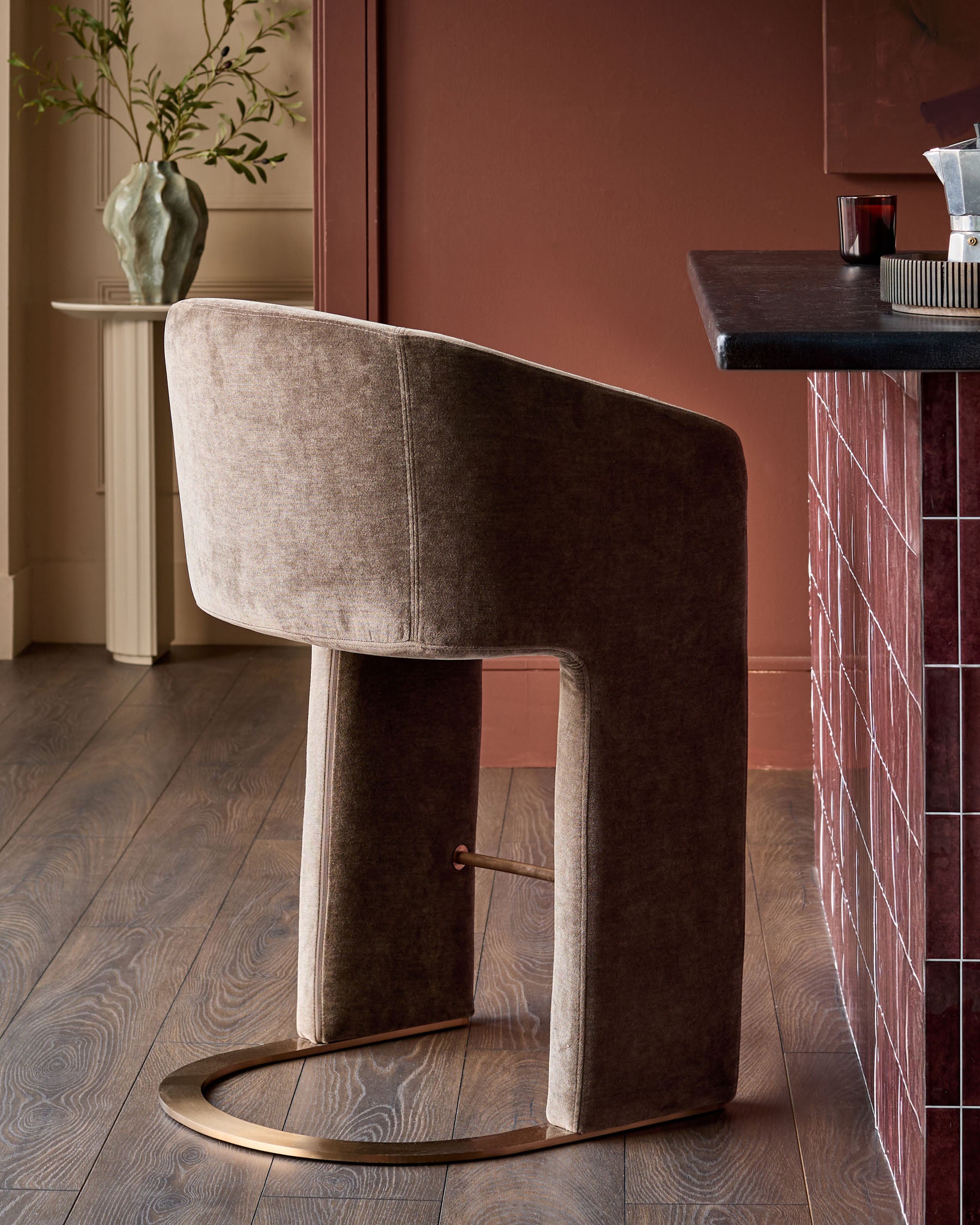 Leighton brushed bronze cantilever bar stool in mocha fabric, shown at angle displaying curved backrest and metal footrest.