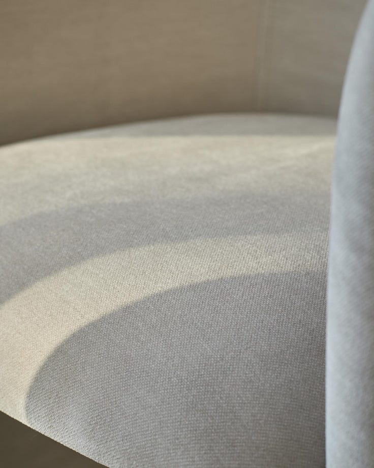 Close-up of a cream coloured upholstered chair seat, with a hint of sunlight and a curved arm.