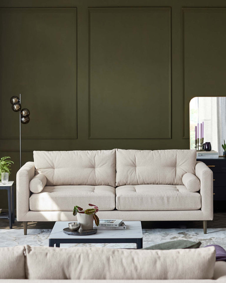 A contemporary three-seater sofa with a neutral beige upholstery and minimalist design, featuring clean lines, plump cushions, and round side pillows. In front of it rests a low-profile rectangular coffee table in a grey finish, supporting a small tray with a decorative plant and candle, as well as a couple of books.