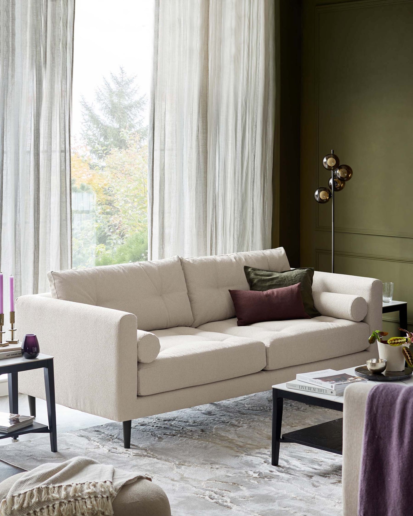 Elegant beige three-seater sofa with plush cushions and clean lines, complemented by a modern black square side table with a lower shelf. The sofa resides on a textured grey area rug, surrounded by soft drapery and a warm, muted room palette.