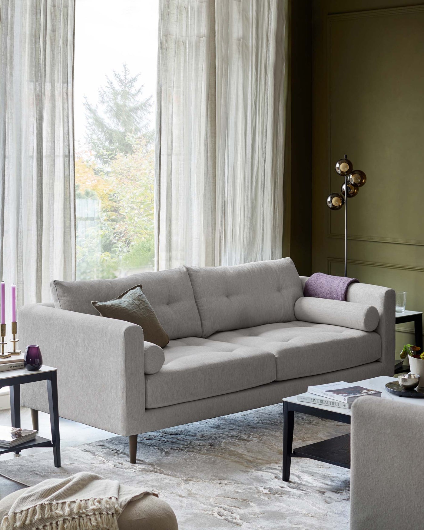 Elegant modern three-seater sofa with plush light grey upholstery and minimalist armrests, complemented by a dark, square-framed side table with a black surface, placed on a textured grey area rug.