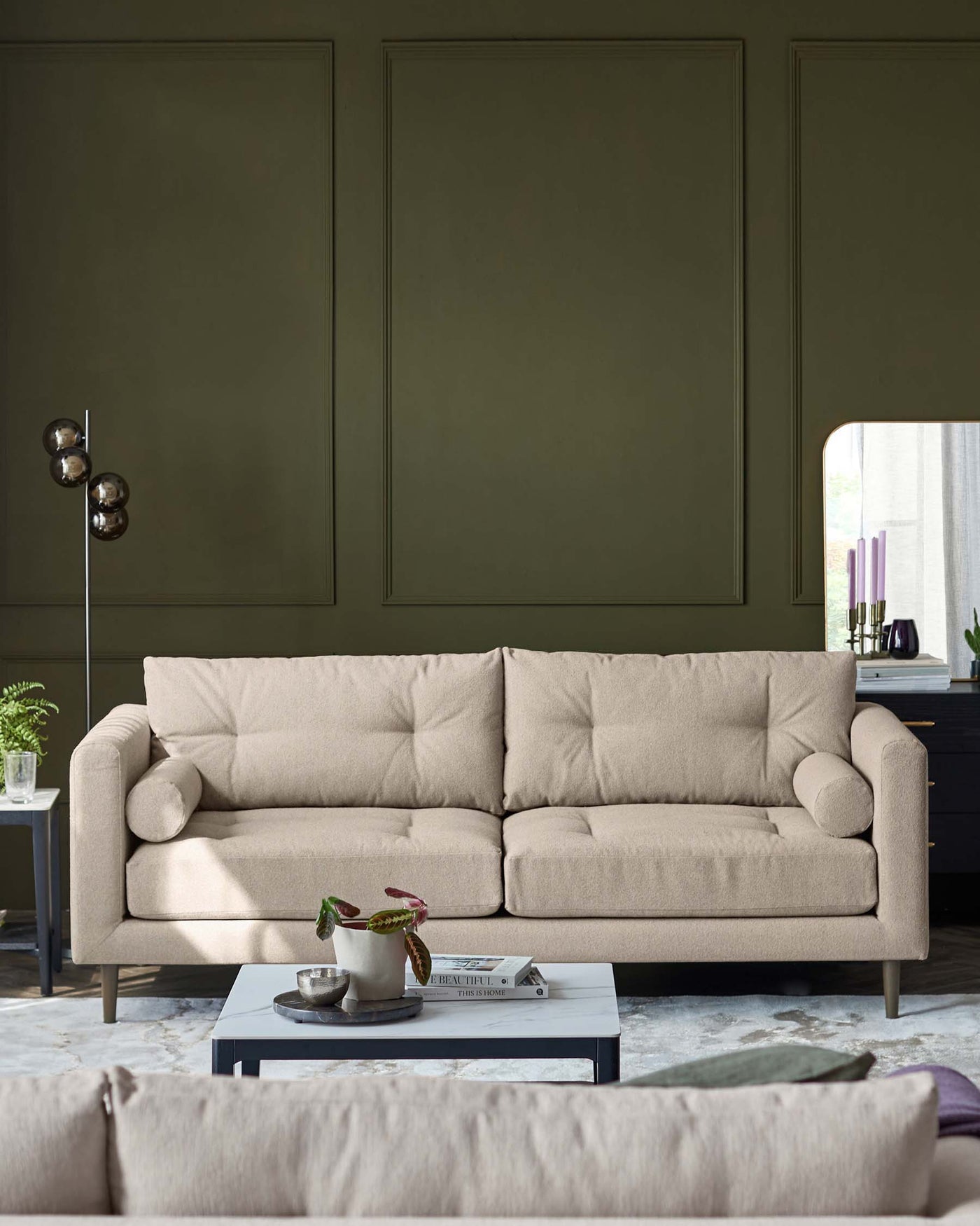 Modern beige three-seater sofa with plush cushions and cylindrical armrests, set in front of an olive green wall, with a low-profile grey coffee table in front, adorned with a plant and decorative items. An off-screen rug is partially visible in the foreground, and a metal floor lamp with round shades stands to the side.