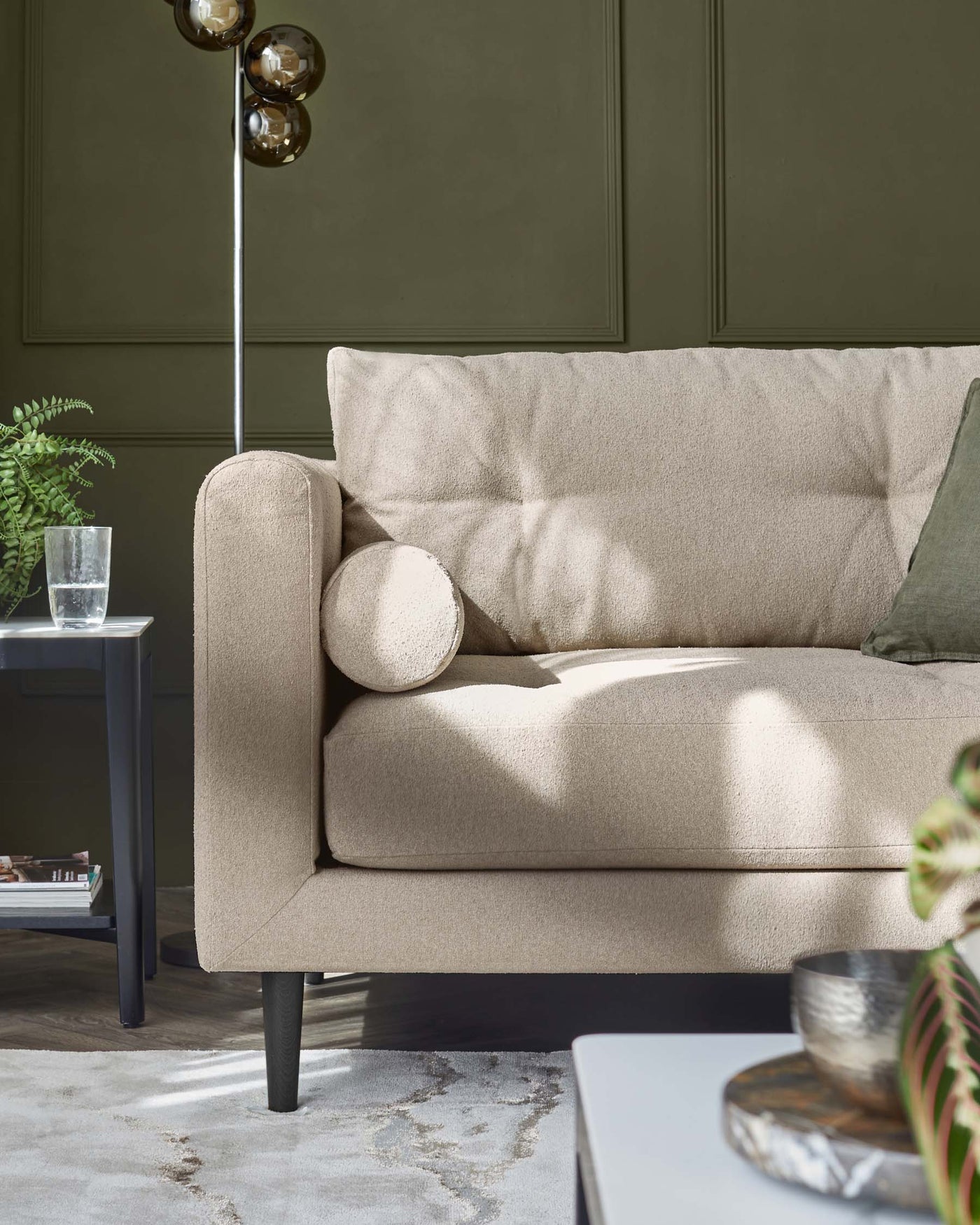 A chic beige fabric sofa with a clean, modern design, featuring cylindrical bolster pillows and minimalist black legs. Next to it is a sleek, black side table with a transparent vase and green foliage on top, complemented by a contemporary-style black coffee table with decorative bowls in the foreground.