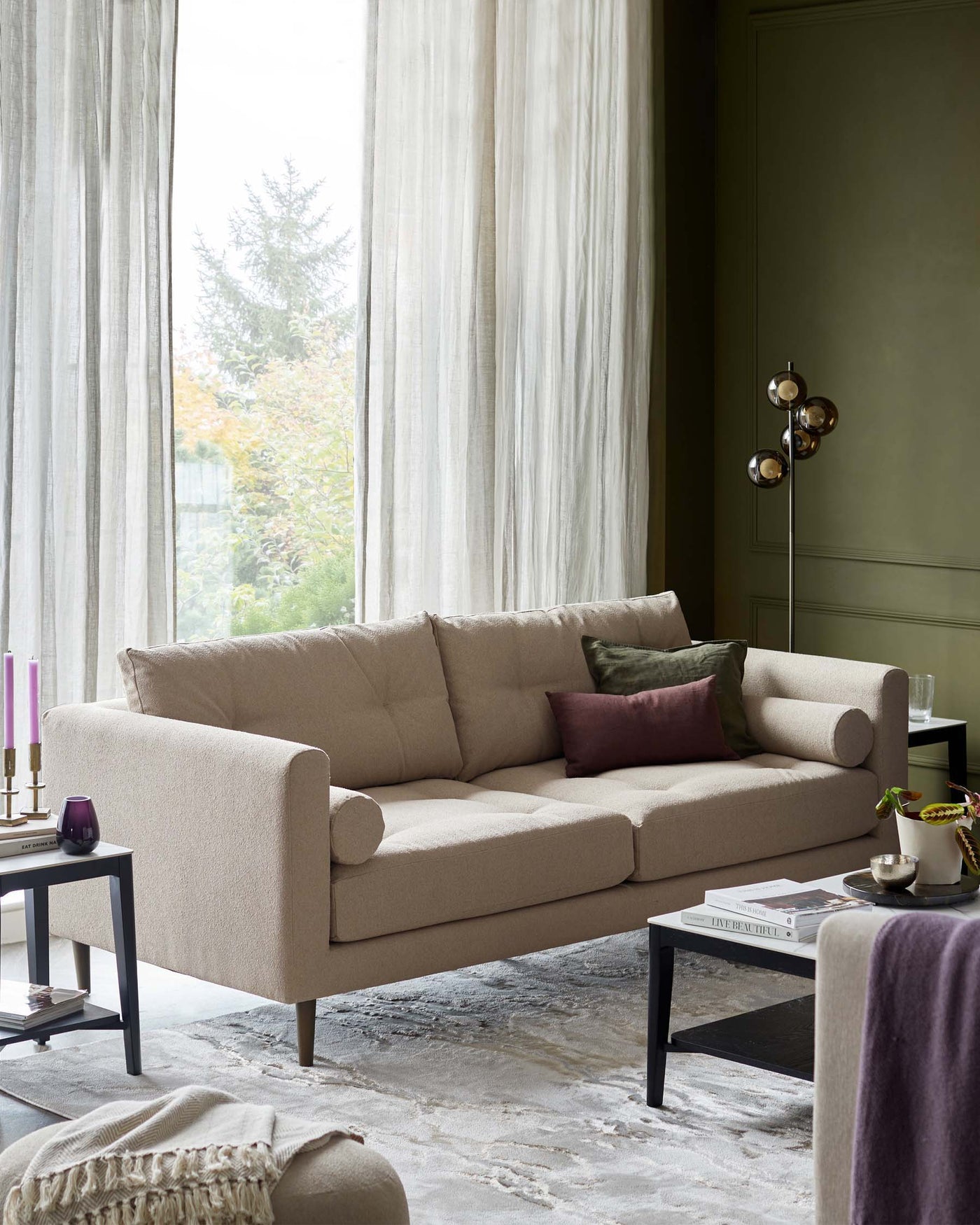 Modern beige three-seater sofa with plush cushions and simple, clean lines, accompanied by a sleek black side table with a square top and thin legs. The setting includes a grey textured area rug and minimalistic decor in a room with muted green walls and large windows draped with sheer curtains.