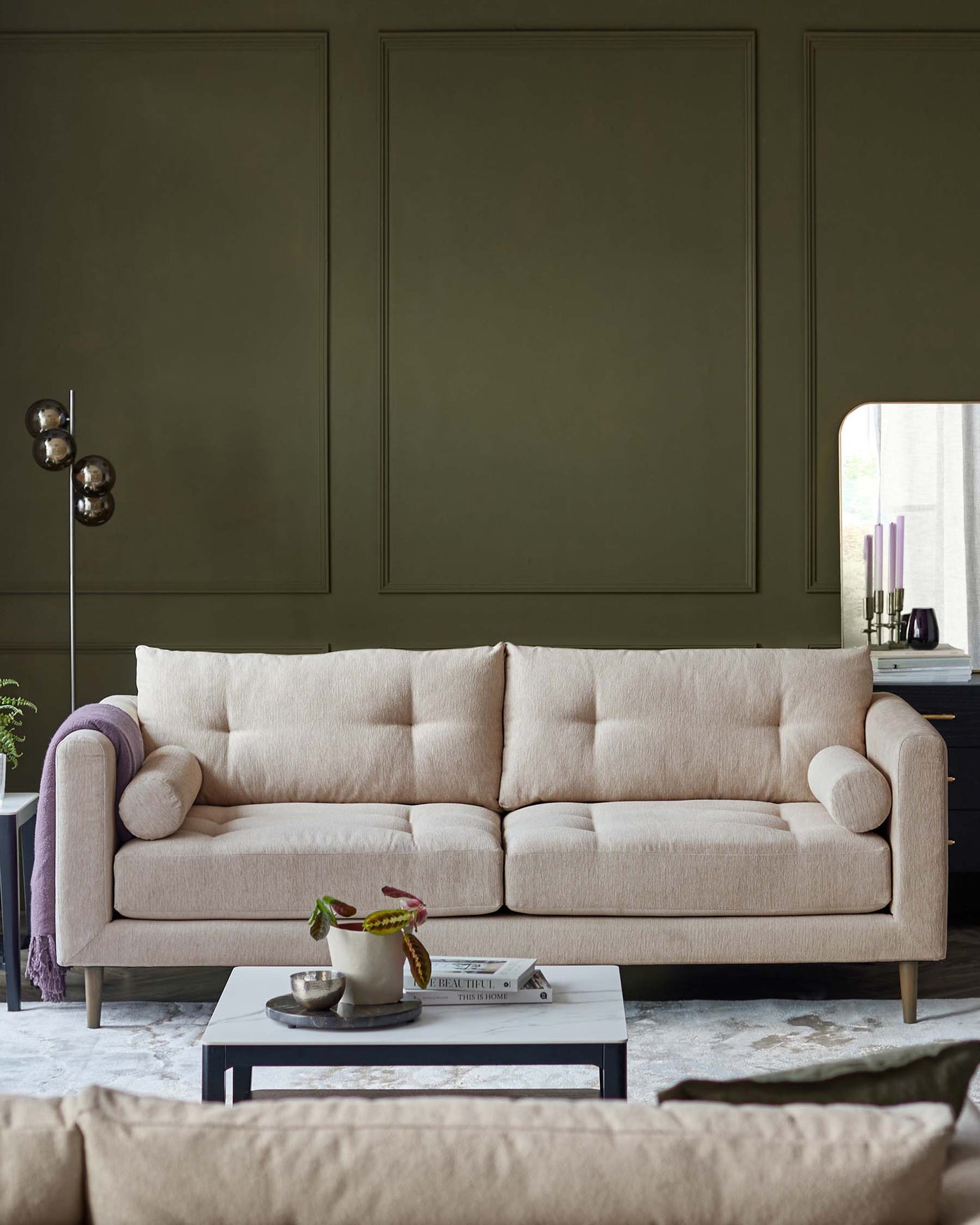 A modern three-seater sofa with a neutral beige fabric upholstery and minimalist design. The sofa features plush back cushions, a single bench seat cushion, and one rounded armrest bolster pillow. It stands on subtle wooden legs and is accompanied by a low-profile, rectangular, grey coffee table with a light-coloured tabletop accentuated with small decorative items and a book.