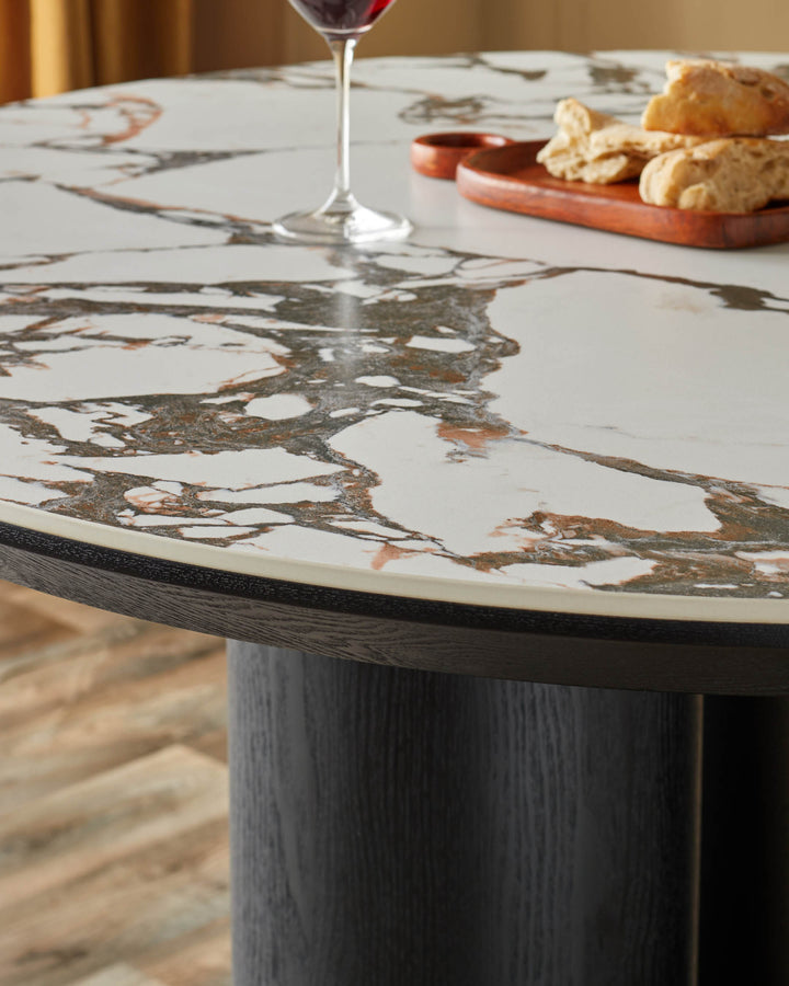 A close up shot of a round dining table with a marble top and a dark wood base, featuring wine and bread on a wooden platter.