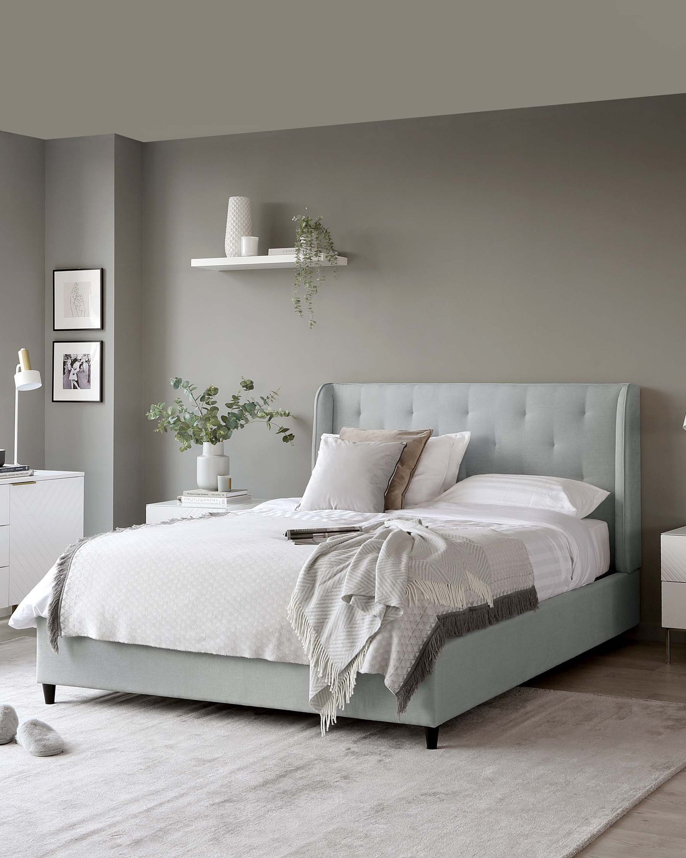 Contemporary bedroom furniture featuring an upholstered queen-size platform bed with a high, tufted headboard in a soft pastel green; matching white bedside tables with a minimalist, modern design; a sleek, white floating wall shelf above the bed displaying decorative items; and a sparse white floor lamp. The ensemble is complemented by soft, neutral bedding and throw blanket, creating a serene and sophisticated space.