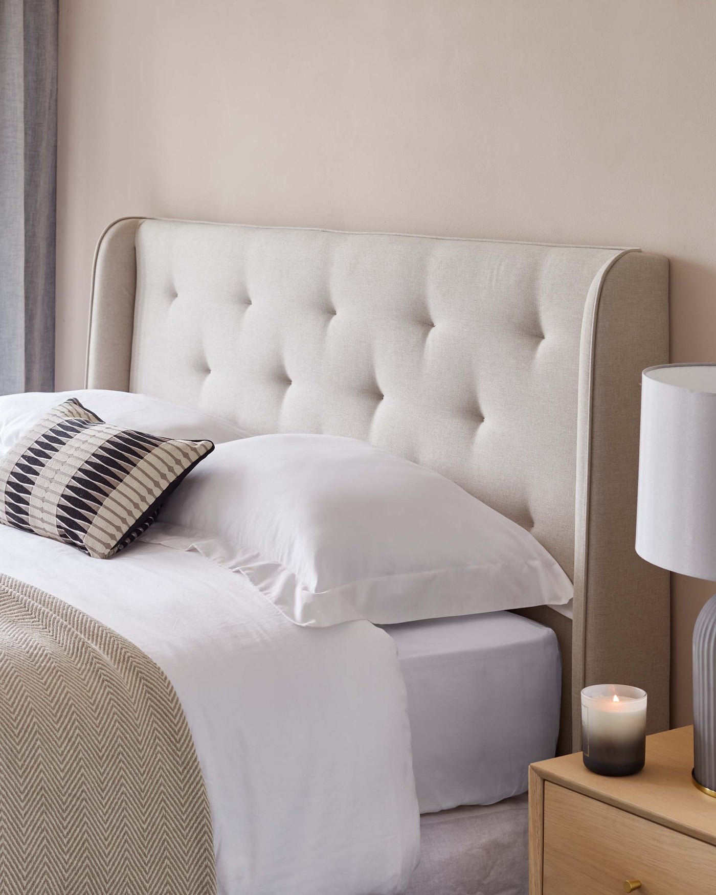 Elegant bedroom scene featuring a light beige, tufted upholstered headboard with a curved top edge, paired with crisp white bedding on a neatly made bed. A wooden bedside table with a smooth finish and round gold handle is placed next to the bed, showcasing a white cylindrical table lamp with a minimalist design and a lite scented candle.